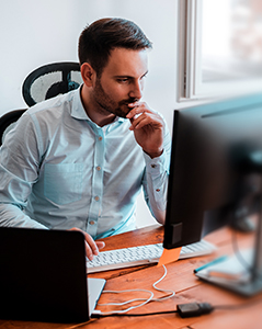 A man in a light blue shirt sits at a desk, focused intently on a computer screen. He rests his hand near his mouth in a thoughtful pose while working with a desktop monitor and laptop. Natural light comes through a nearby window, illuminating the workspace.