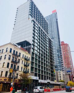 Street-level view of a high-rise building under construction in an urban setting, surrounded by completed buildings and city traffic, showcasing vertical development in a dense metropolitan area.