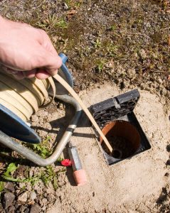 A person feeds a flexible hose or cable into an underground pipe access point, possibly for inspection or environmental sampling. The equipment includes a reel for the hose, and the surrounding area is a mix of soil and sparse vegetation.