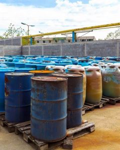 Rows of weathered metal drums, some rusted, are stored on wooden pallets in an outdoor industrial area. The barrels are blue and brown in color, suggesting possible storage of hazardous materials or chemicals. A concrete wall and industrial piping are visible in the background.