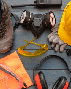 Collection of personal protective equipment (PPE) arranged on a wooden surface, including a hard hat, safety goggles, work boots, gloves, respirator mask, safety glasses, ear protection, and a high-visibility vest.
