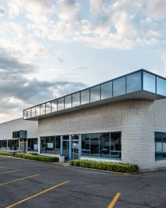 Modern commercial or industrial building with large windows and a prominent glass-wrapped second-story overhang. The structure is surrounded by an empty parking lot with clearly marked spaces, under a partly cloudy sky.
