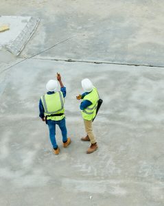 Two construction workers wearing safety vests, hard hats, and work boots stand on a concrete surface. One is pointing upward while the other looks in the same direction, possibly discussing site details or inspecting the structure above.