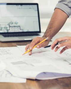 lose-up of two people reviewing environmental and construction drawings spread out on a wooden table. One person is holding a pencil while the other gestures with a pen. In the background, a laptop displays a digital floor plan, suggesting a design or construction planning session.