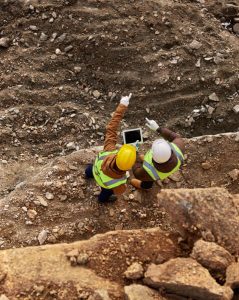 Two construction workers wearing safety vests and hard hats stand at the edge of a rocky excavation site. One holds a tablet while the other points toward an area of interest, indicating they are discussing or inspecting the site.