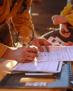 Worker in a yellow protective uniform filling out safety or inspection forms on a clipboard outdoors, with another person in similar attire visible in the background, suggesting a field safety or compliance check.