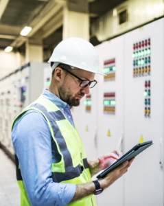 Energy auditor in a hard hat, holding a tablet, taking consumption measurements in a control room.