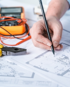 Close-up of a person marking a technical blueprint with a pen, surrounded by electrical tools including a multimeter and pliers, representing detailed planning and precision in engineering or construction projects.